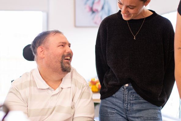 A man sits in a wheelchair in his home, smiling up at other people