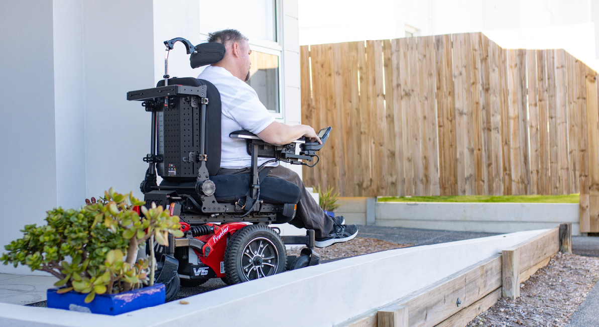 A man in a powered wheelchair is driving down an accessible pathway situated at the front of his home.