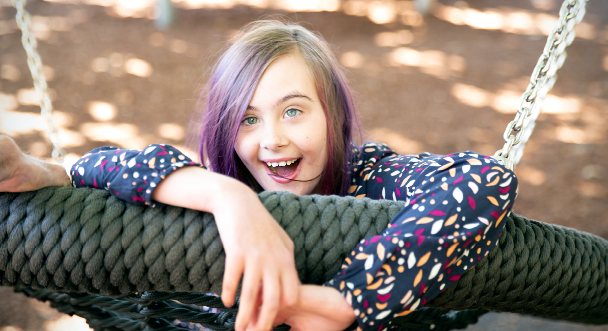 A girl with purple hair is being pushed on big swing and smiling at the camera