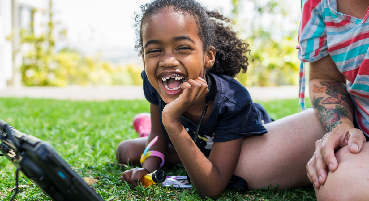 A girl is laying on her stomach on the grass with a tablet and someone beside her. She is smiling at the camera.