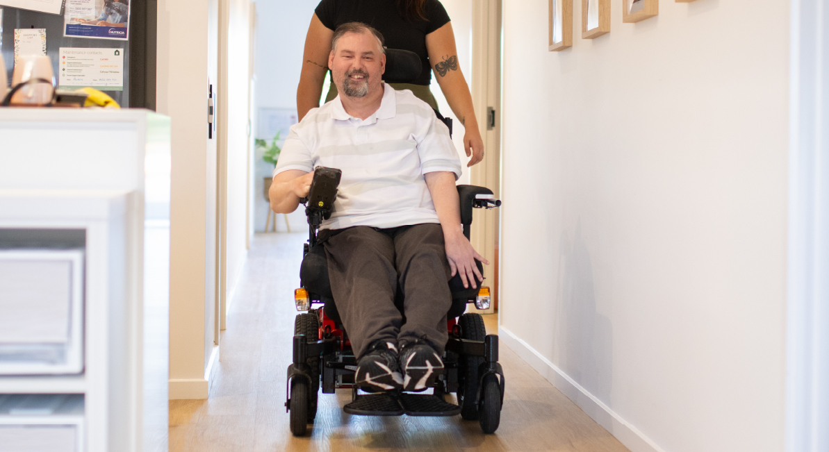 A man in a wheelchair is rolling down the hallway of a SIL home with a support worker walking behind him. He is smiling at the camera.